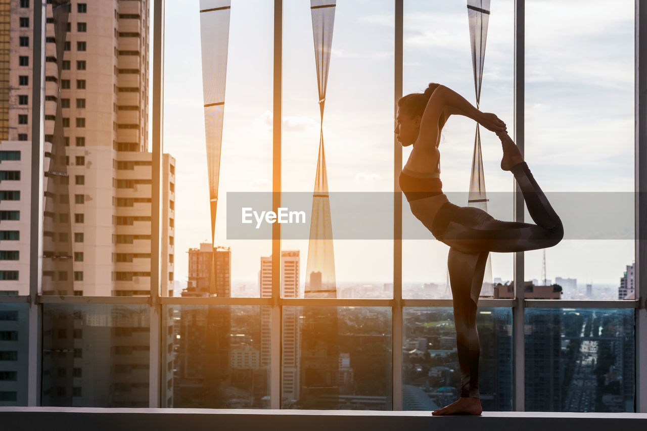 Side view of young woman exercising by window against cityscape