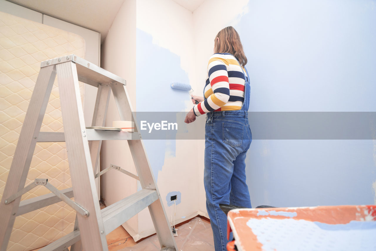 Standing woman dressed in overalls and striped blouse, seen from behind, painting a white wall