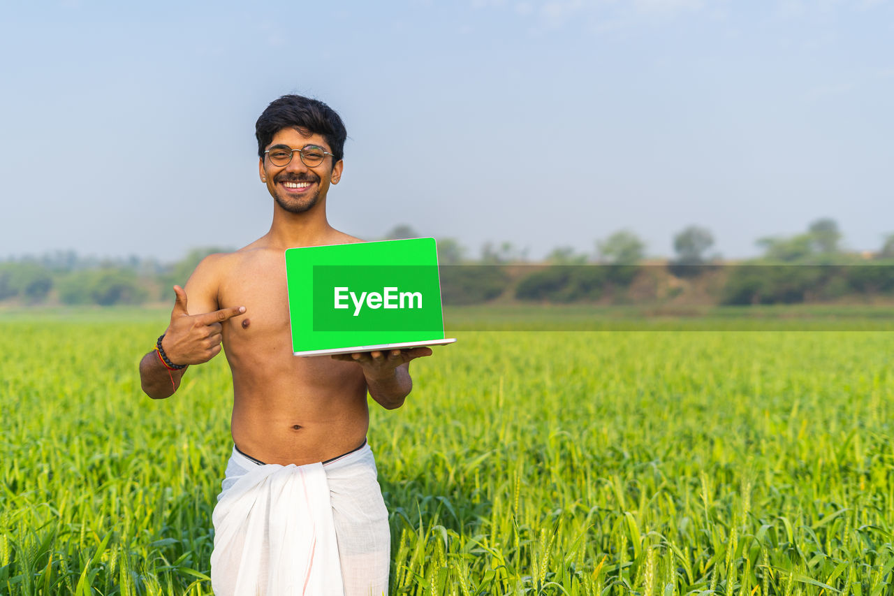 agriculture, one person, field, paddy field, crop, adult, plant, rural scene, land, nature, green, smiling, grass, landscape, grassland, men, environment, yellow, technology, sky, happiness, meadow, rural area, standing, portrait, emotion, communication, copy space, lawn, young adult, person, wireless technology, flower, outdoors, growth, summer, occupation, holding, three quarter length, cheerful, looking at camera, day, glasses, front view, environmental conservation, computer, farm, fashion