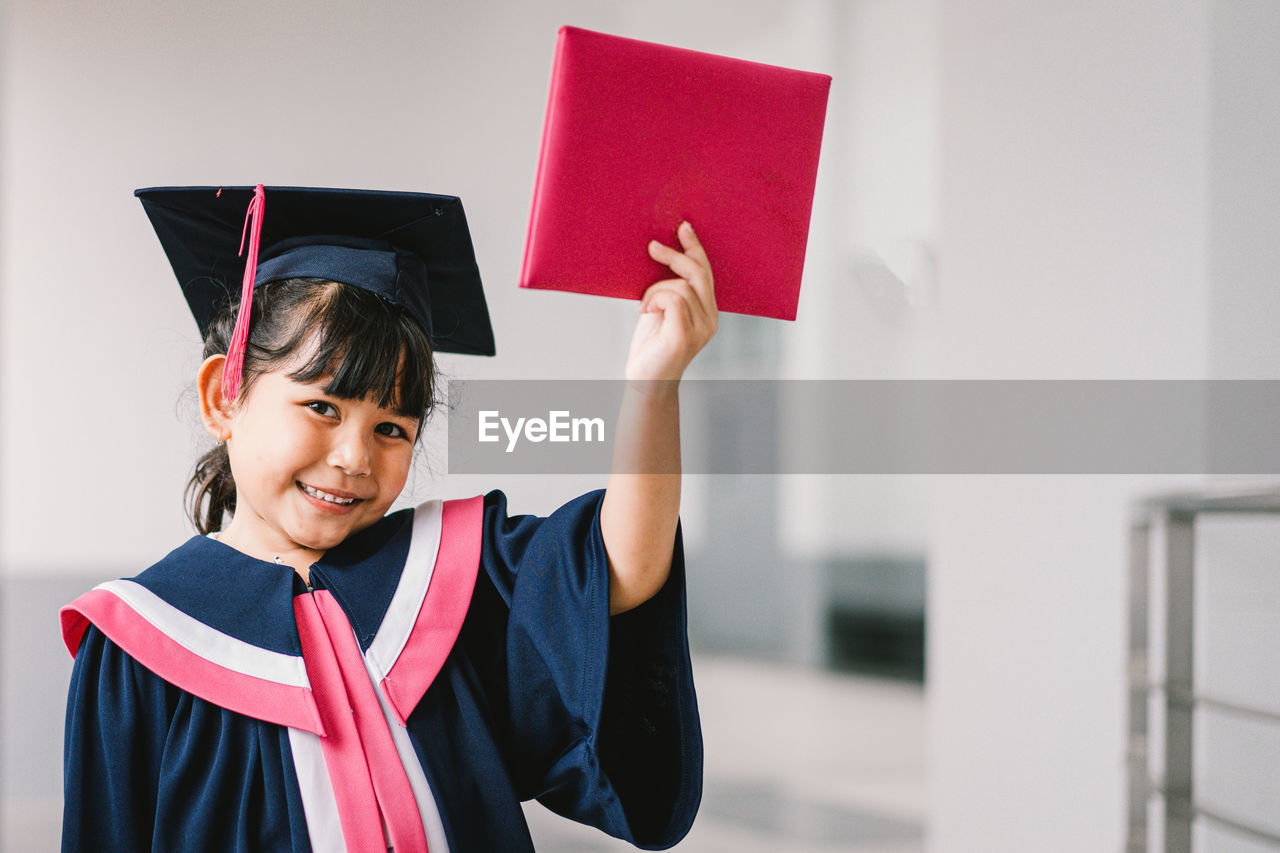 graduation, achievement, graduation gown, one person, happiness, academic dress, smiling, student, emotion, education, mortarboard, success, university, women, school, portrait, cheerful, holding, learning, university student, adult, positive emotion, indoors, copy space, female, young adult, pride, child, standing, event, aspirations, celebration, looking at camera, certificate, childhood, diploma, front view