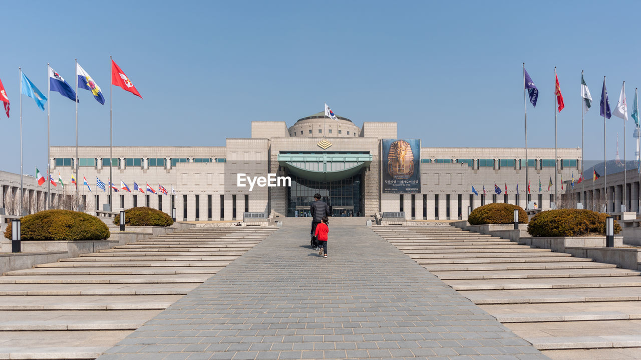 walkway, architecture, flag, boardwalk, patriotism, sky, built structure, travel destinations, building exterior, nature, clear sky, city, history, town square, the past, travel, government, landmark, sunny, day, outdoors, tourism, blue, building, footpath, plaza, adult, full length