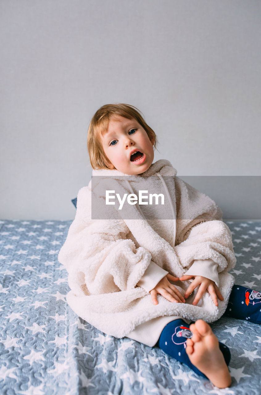 Cute child y on the bed in cozy adult clothes and a hat with earflaps.