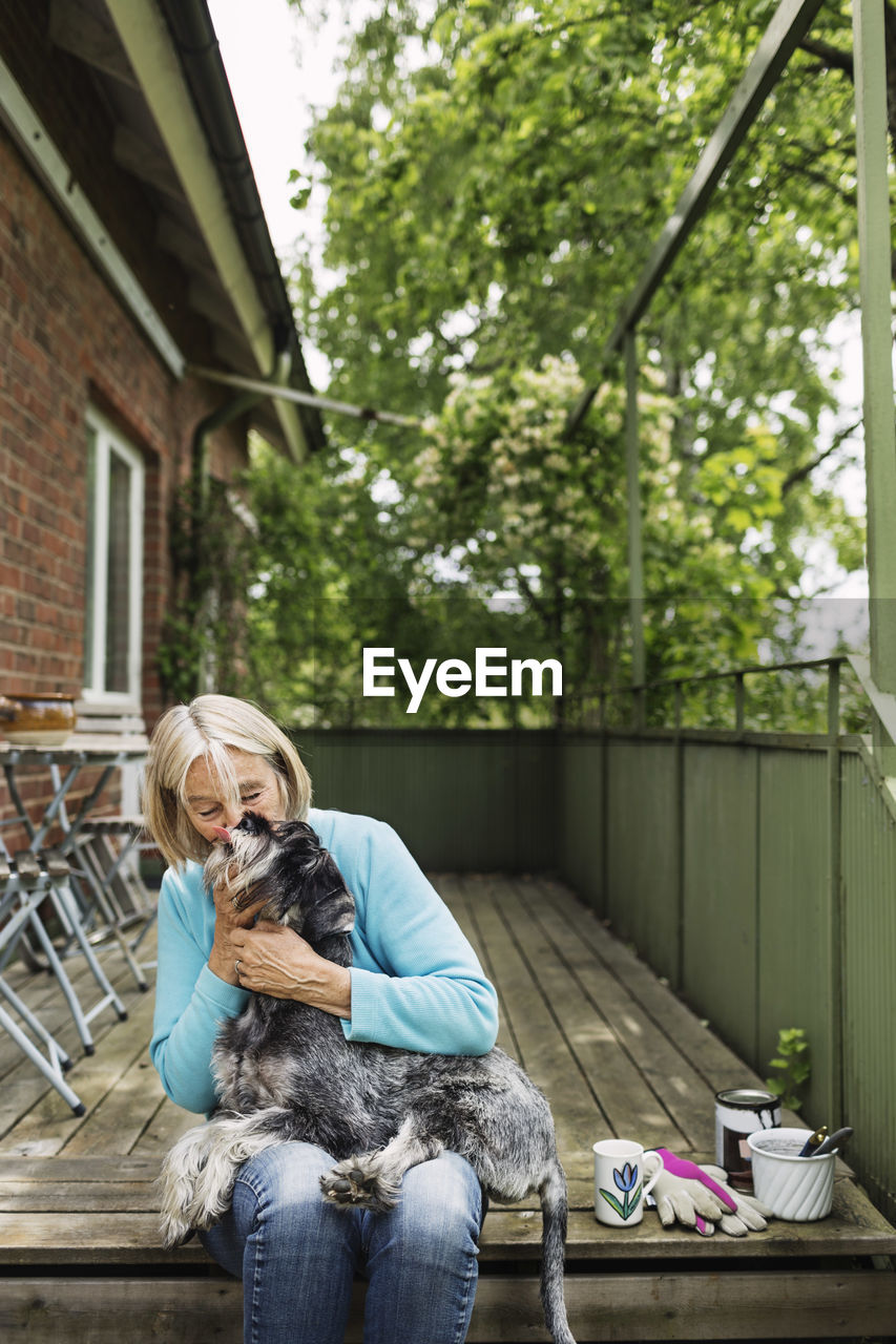 Happy senior woman kissing dog while sitting on porch