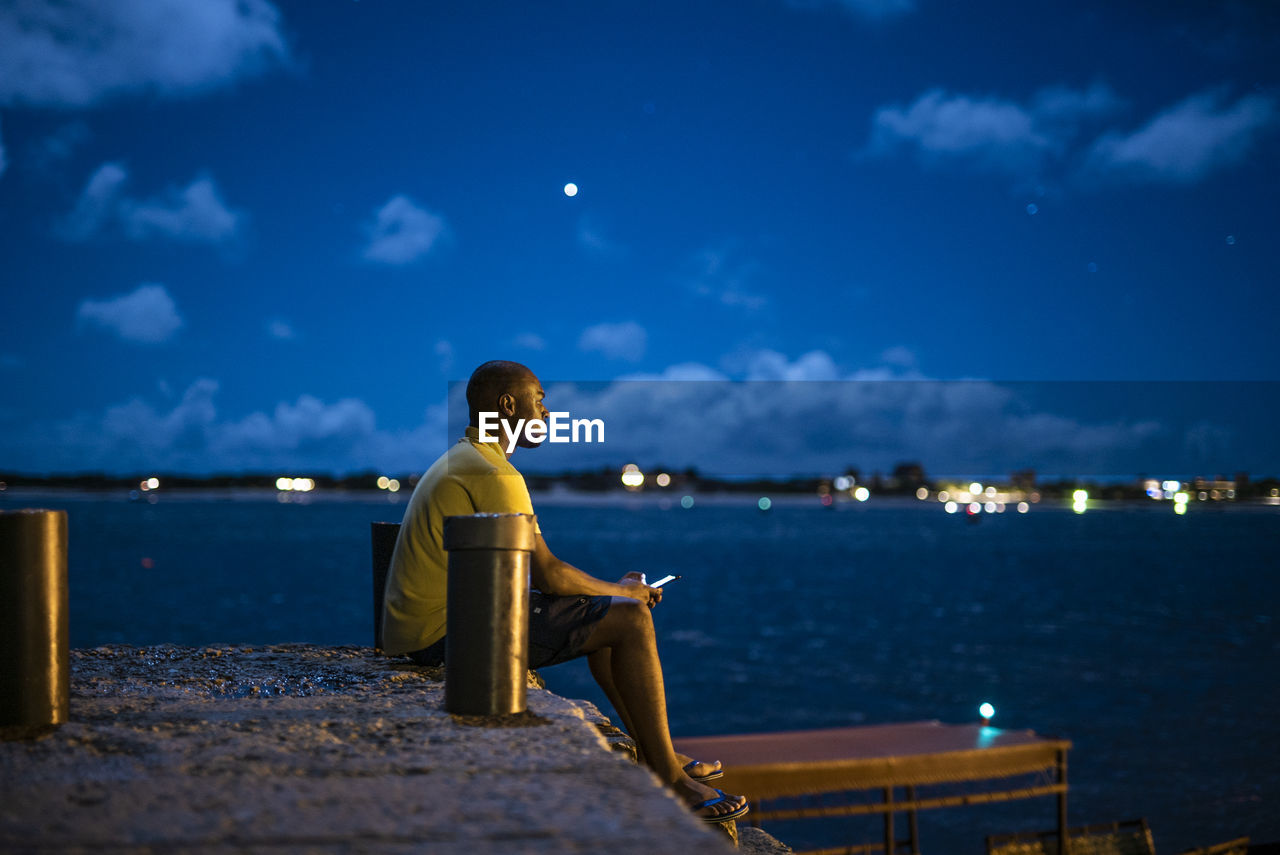 Side view of man sitting on pier by sea at night