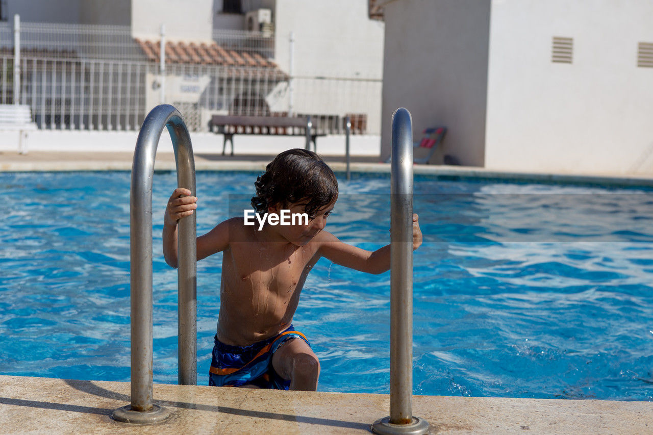 A young boy grips a ladder while getting out of a sunlit pool.