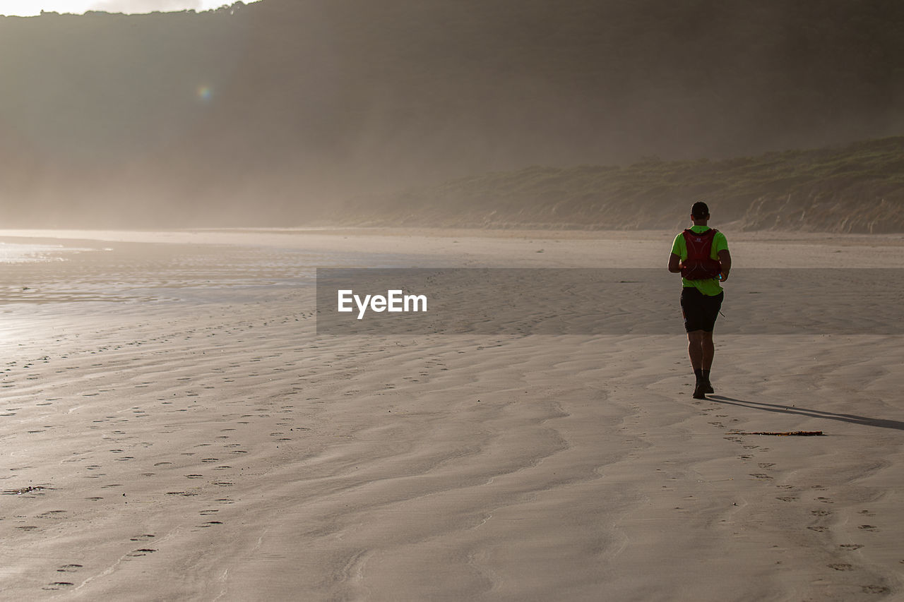 REAR VIEW OF MAN WALKING ON BEACH AGAINST SEA