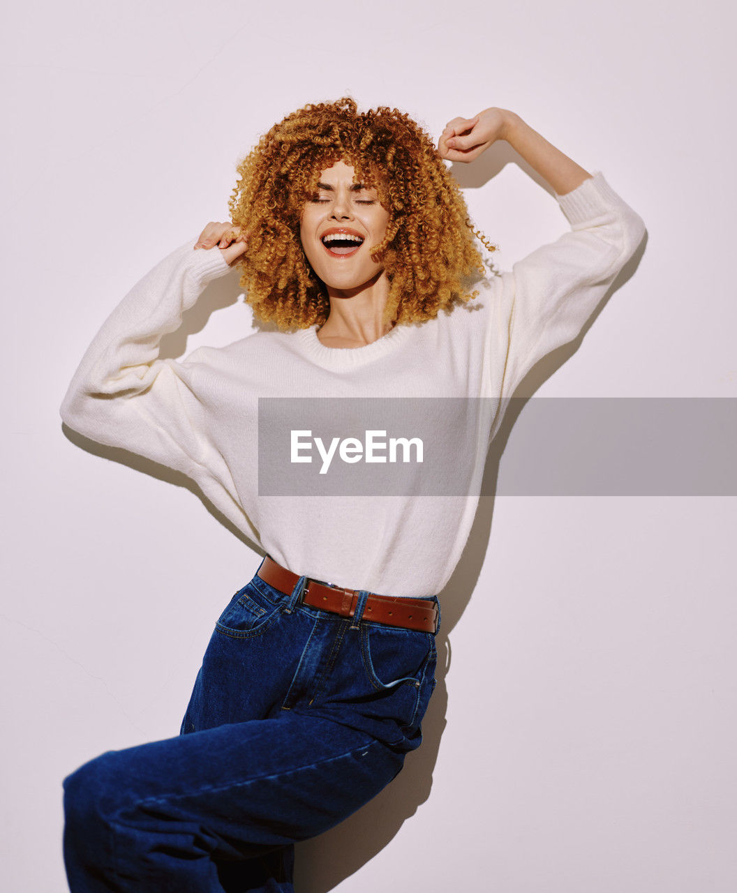 studio shot, one person, curly hair, adult, portrait, women, indoors, jeans, photo shoot, young adult, hairstyle, clothing, redhead, trousers, casual clothing, looking at camera, emotion, female, person, three quarter length, smiling, fashion, sleeve, long hair, happiness, outerwear, textile, facial expression, white background, limb, positive emotion, mouth open, arm, front view, human hair, fun, vitality, pocket, human mouth, standing, relaxation, copy space, brown hair