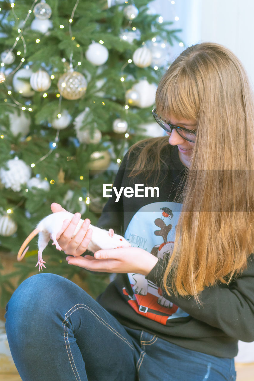 Girl holding rat while sitting against christmas tree