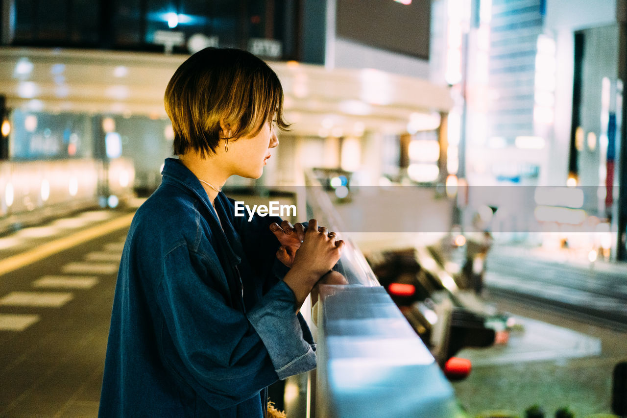 YOUNG WOMAN LOOKING AT CAMERA WHILE STANDING AT ILLUMINATED BUS