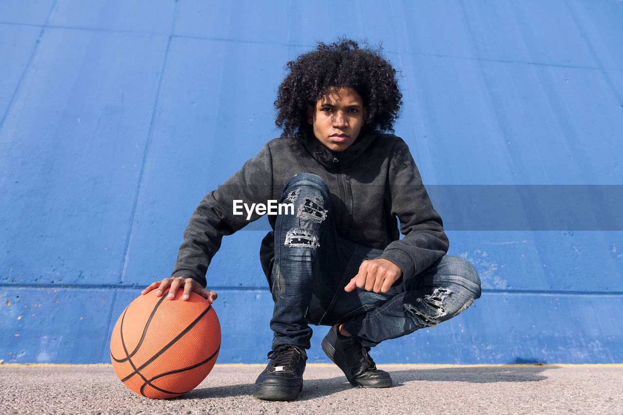 Full body of serious african american teen male in informal outfit sitting with basketball ball near blue wall and looking at camera