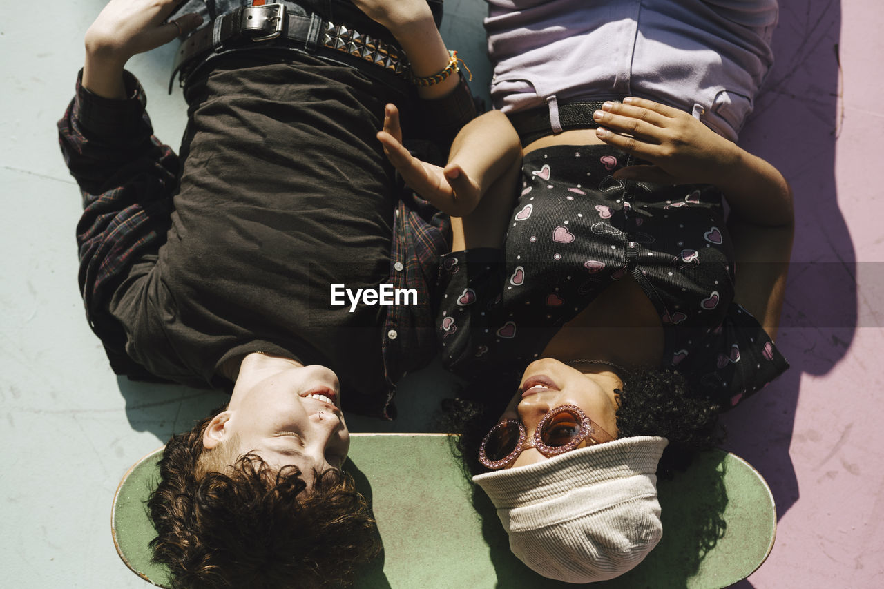 Smiling teenage boy and girl lying on skateboard over footpath during sunny day