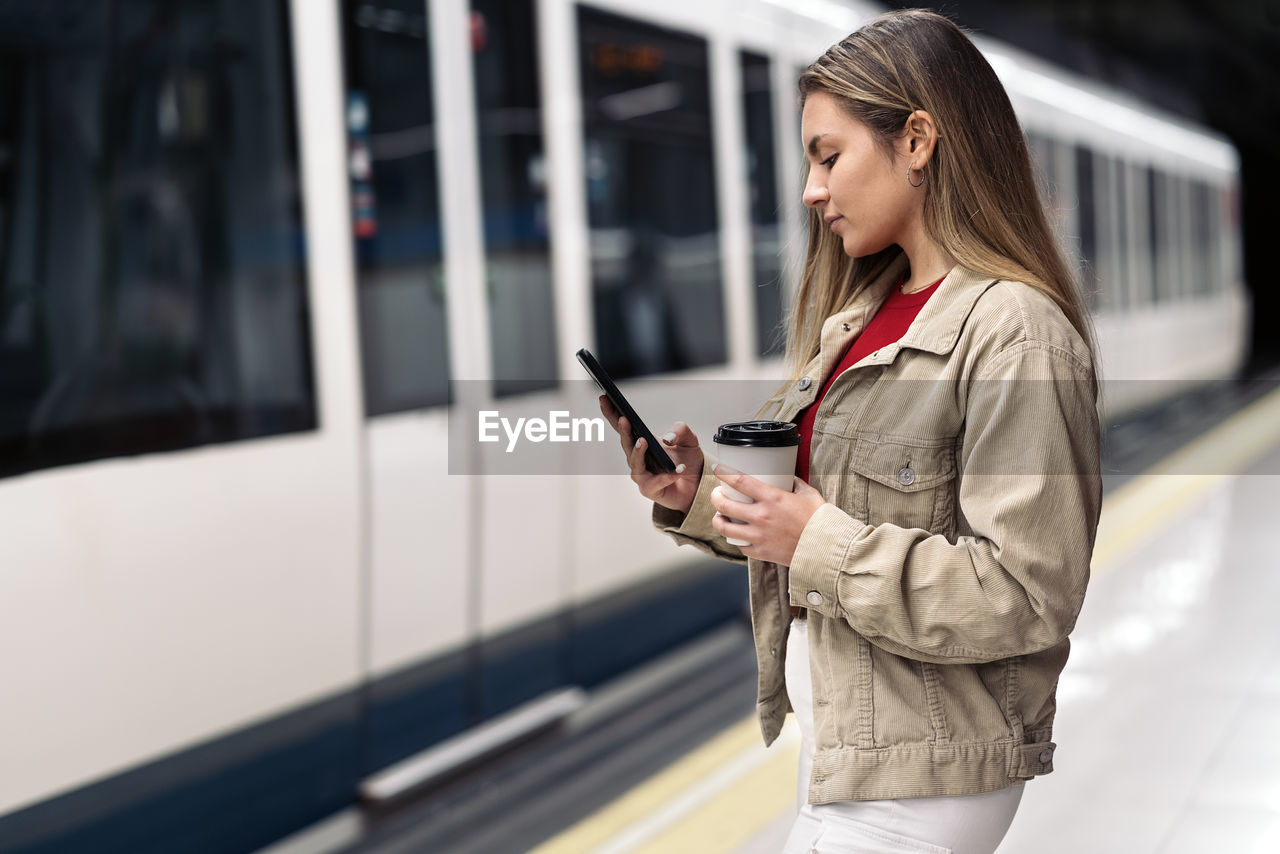 FULL LENGTH OF YOUNG WOMAN USING PHONE WHILE STANDING ON MOBILE