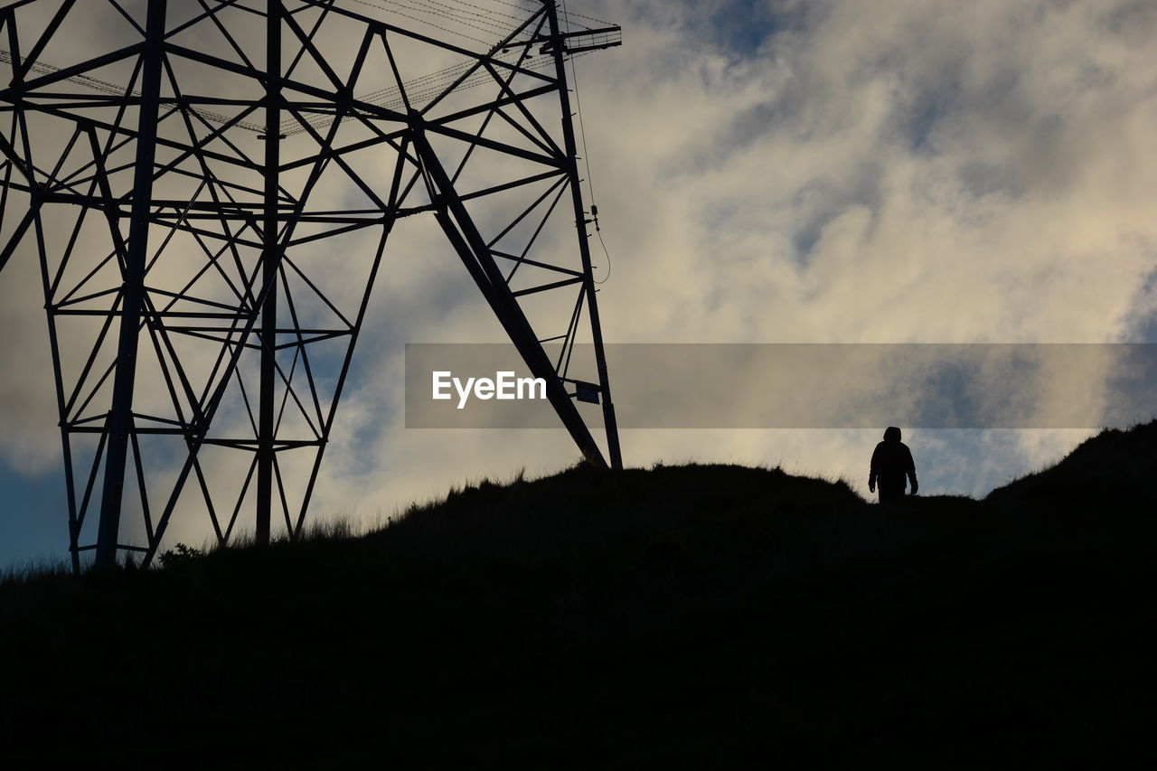 Low angle view of silhouette man standing by electricity pylon against cloudy sky