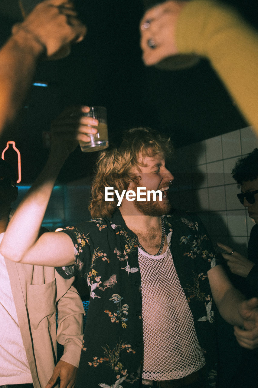 Cheerful young man holding drink dancing with friends at nightclub