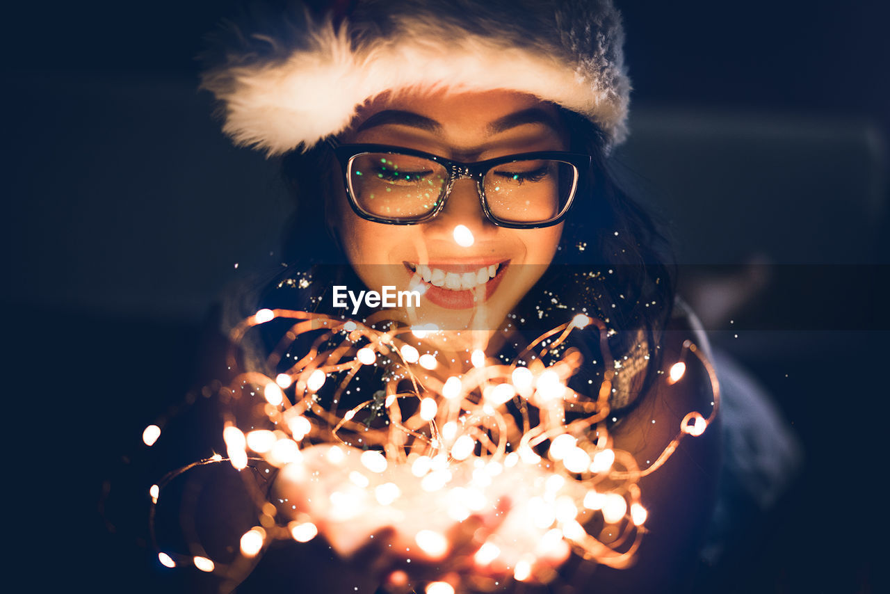 Close-up of smiling young woman with illuminated string lights in darkroom