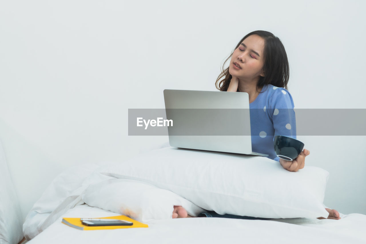 YOUNG WOMAN USING PHONE WHILE SITTING ON WHITE WALL