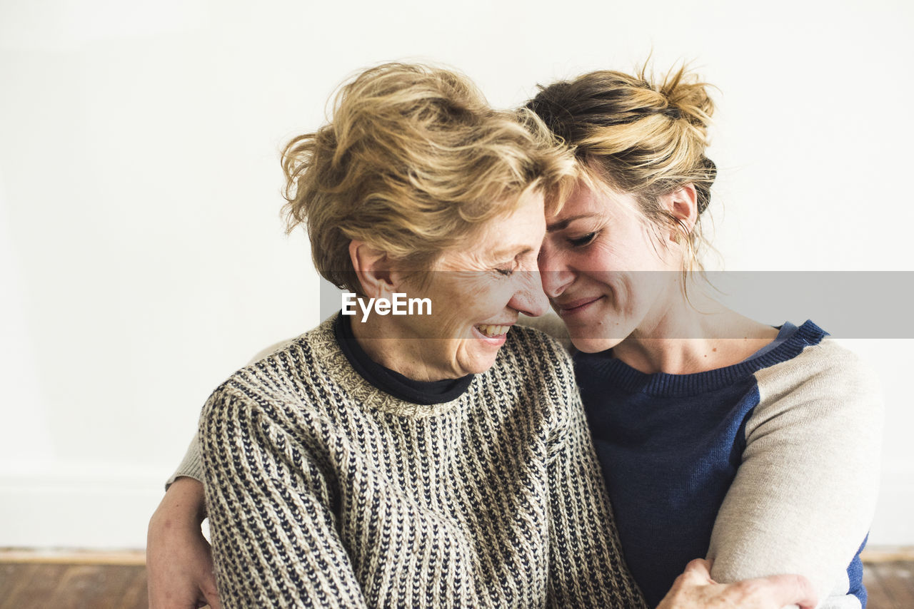 Close-up of smiling mother and daughter sitting against wall at home