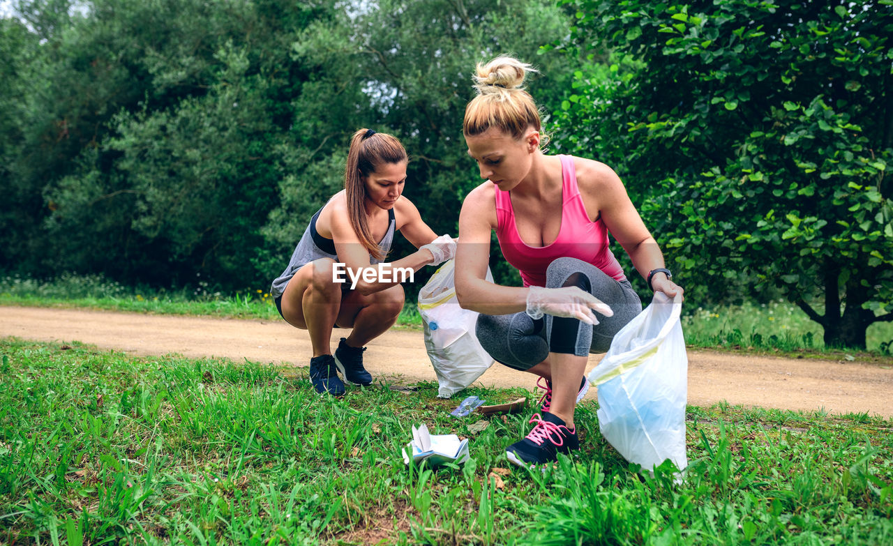 Full length of happy women picking up garbage while crouching on grass against trees