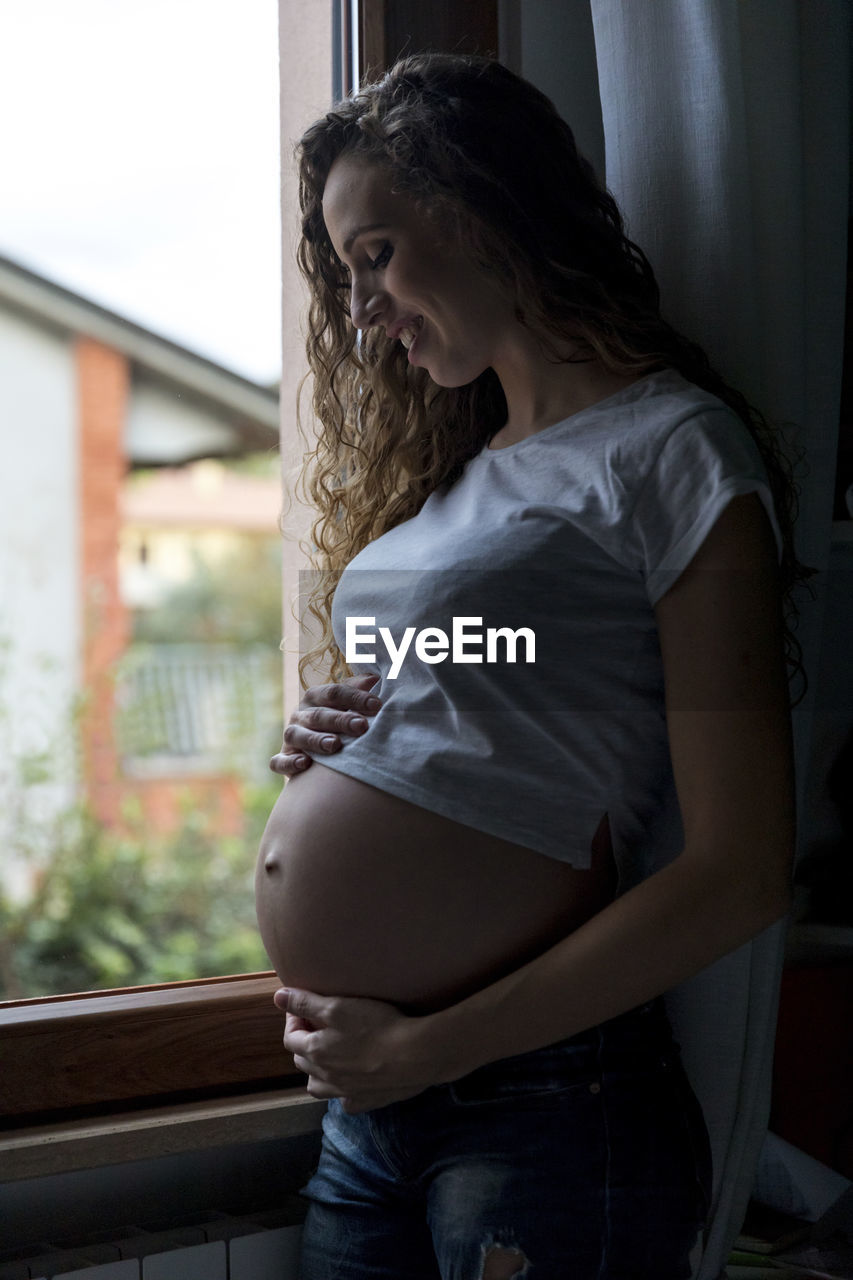 Smiling pregnant woman standing with hands on stomach at home