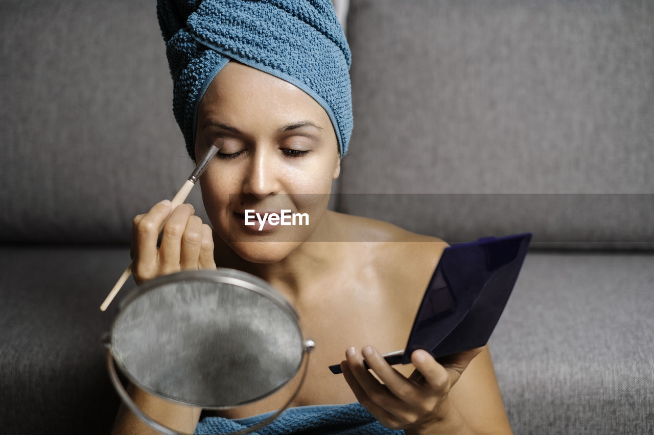 Content young bare shouldered female with towel on head applying eye shadow on eyebrows during makeup procedure after shower at home