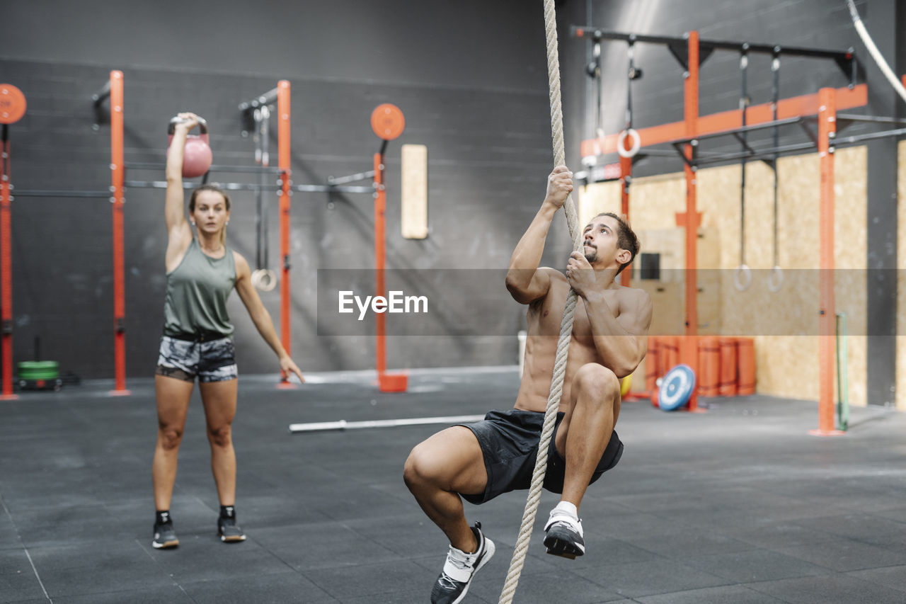 Young man exercising with rope while girlfriend lifting kettlebell in gym