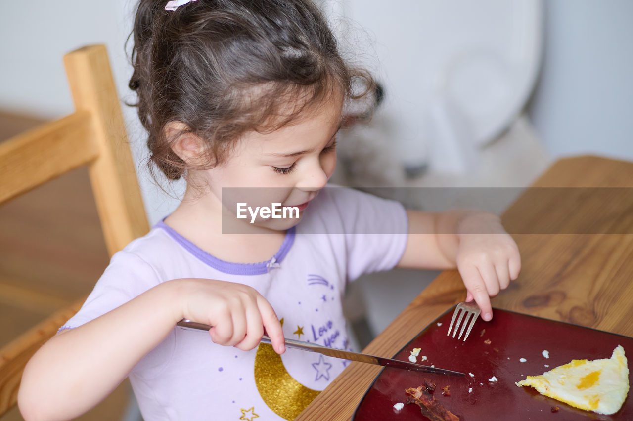 Young girl eating eggs and bacon with knife and fork for the first time