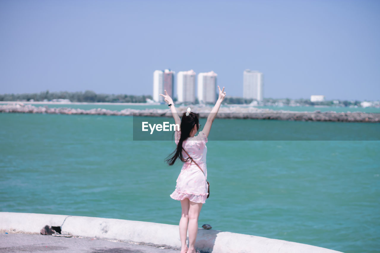 Rear view of woman with arms raised standing against sea in city