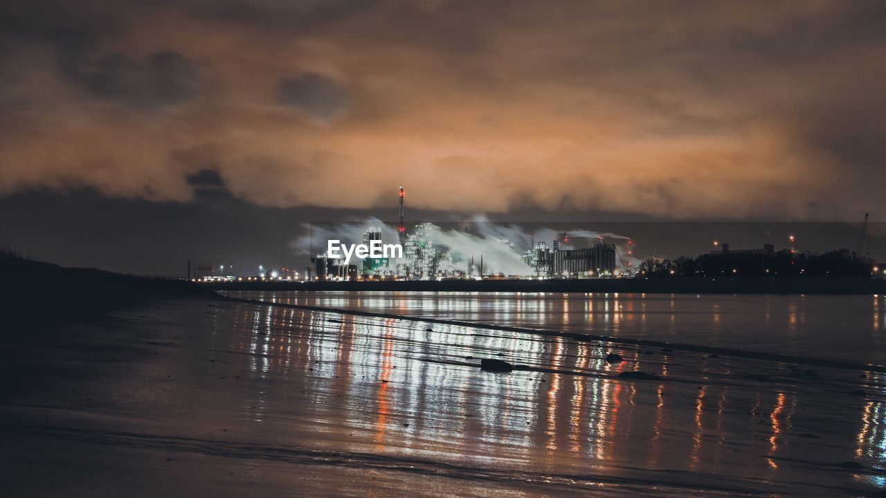 Illuminated factory by river against cloudy sky at night