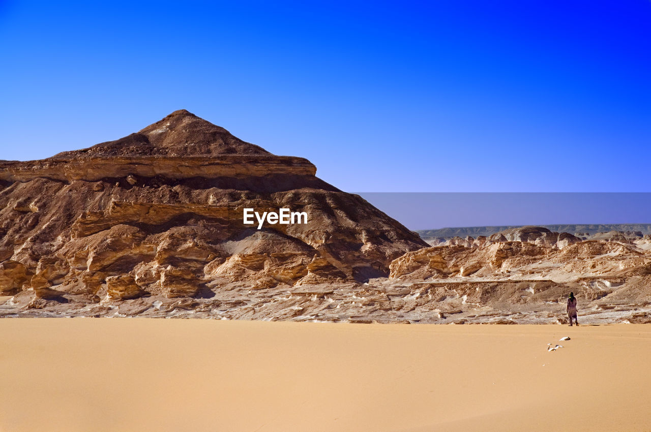 Distant view of man standing by rock formations against clear blue sky