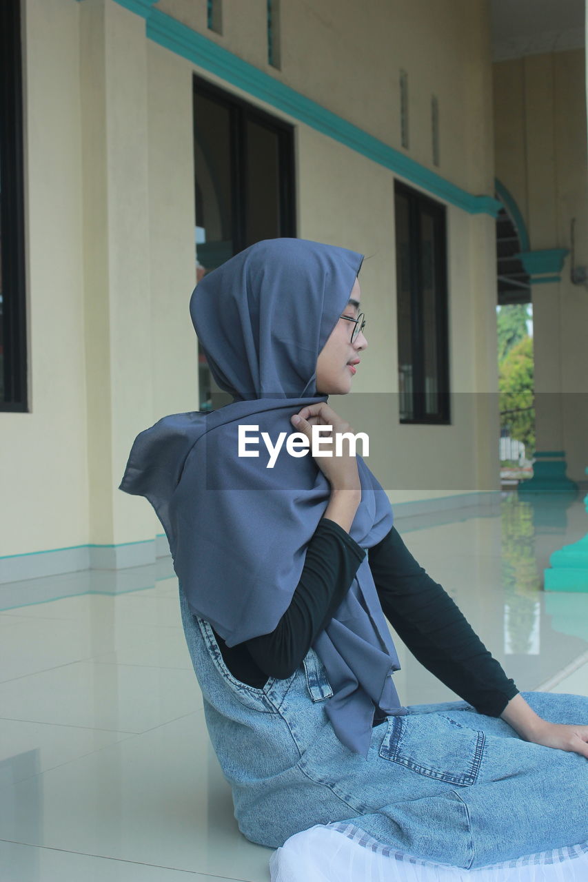 Young woman wearing hijab sitting against wall