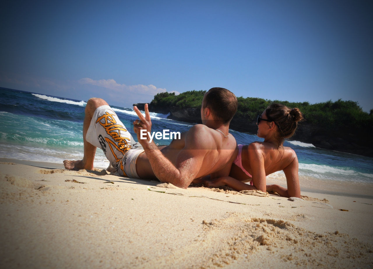 Tilt image of couple resting on beach against blue sky during sunny day