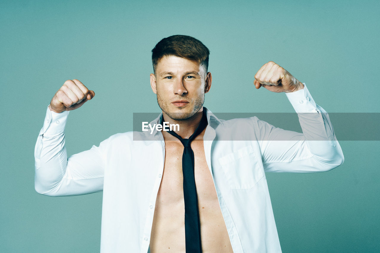 one person, adult, men, studio shot, waist up, colored background, formal wear, person, clothing, portrait, indoors, arm, young adult, standing, front view, sports, button down shirt, gesturing, shirt, businessman, blue background, arms raised, athlete, human limb, blue, limb, business, human face, looking at camera, white, necktie, menswear, lifestyles, occupation, shirt and tie, exercising, muscular build, green background