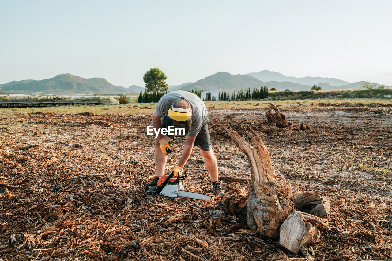 Mature unrecognizable man standing in field and starting modern chainsaw while getting ready for cutting tree trunk in rural area in highlands