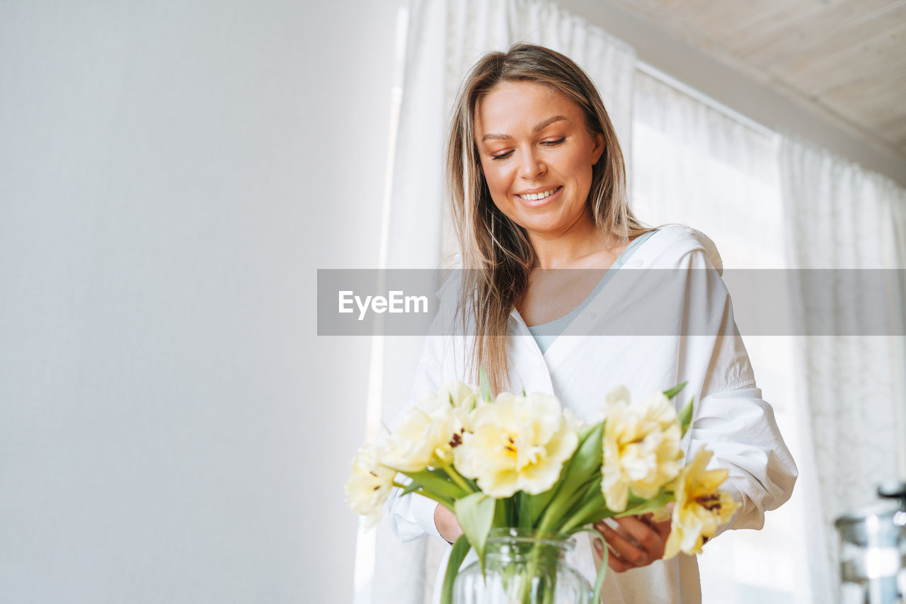 Young smiling woman with blonde long hair in white shirt with bouquet of yellow flowers at home