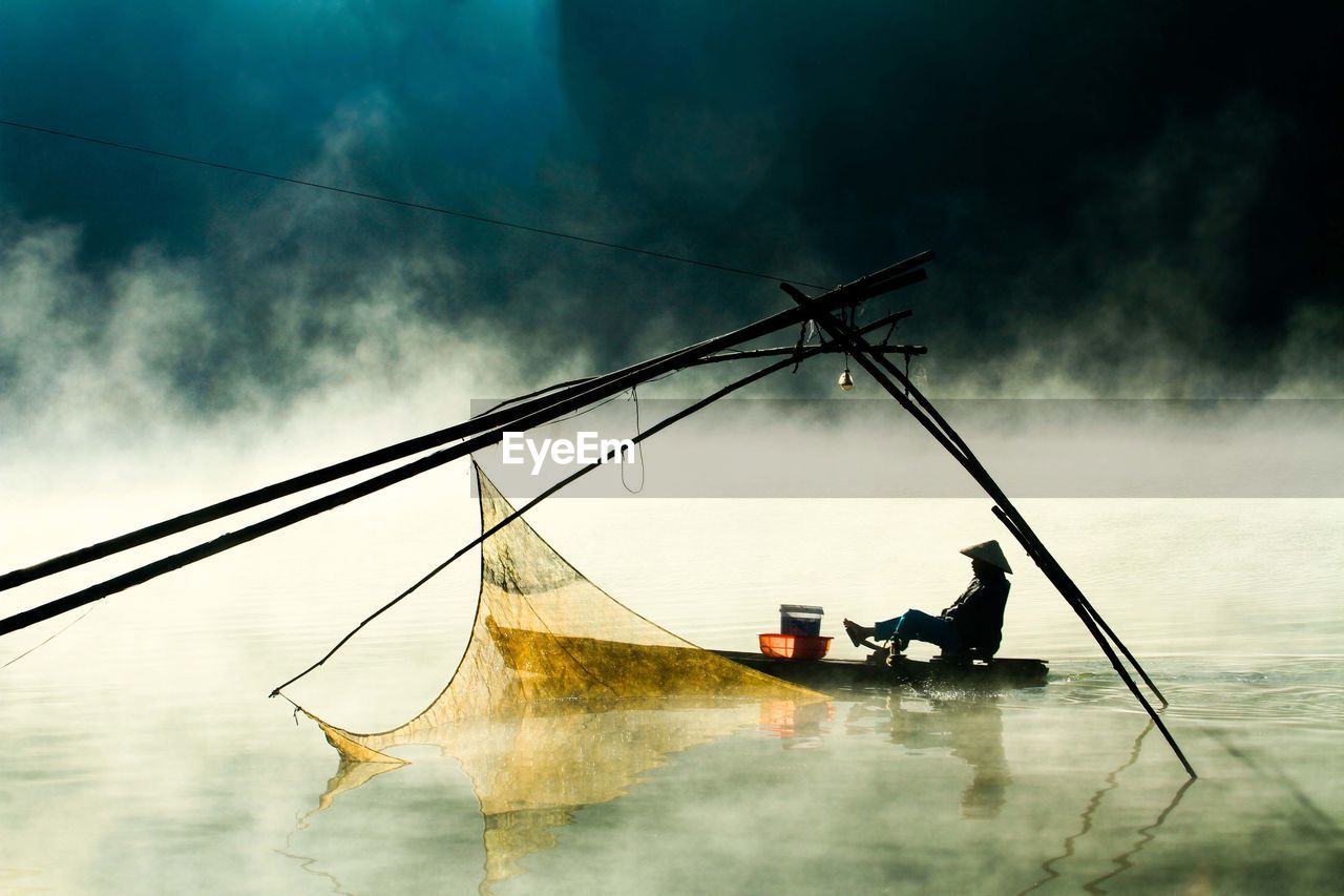 Man in boat on lake by fishing net during foggy weather