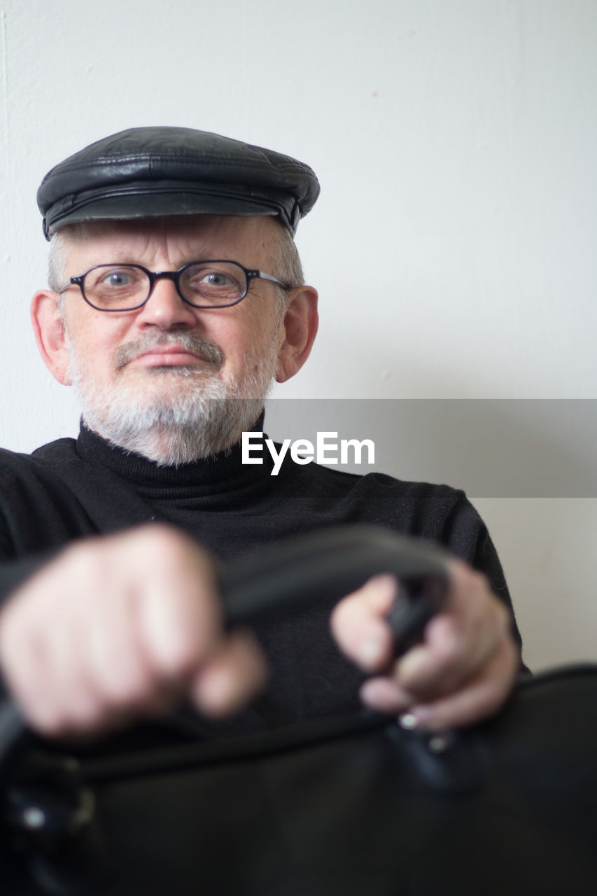 Portrait of senior man wearing flat cap and eyeglasses while holding briefcase at home