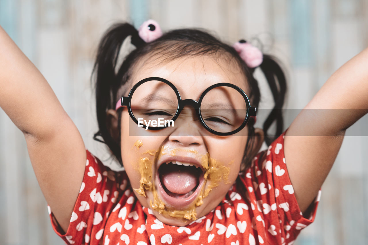 Close-up portrait of cheerful girl eating peanut butter against wall