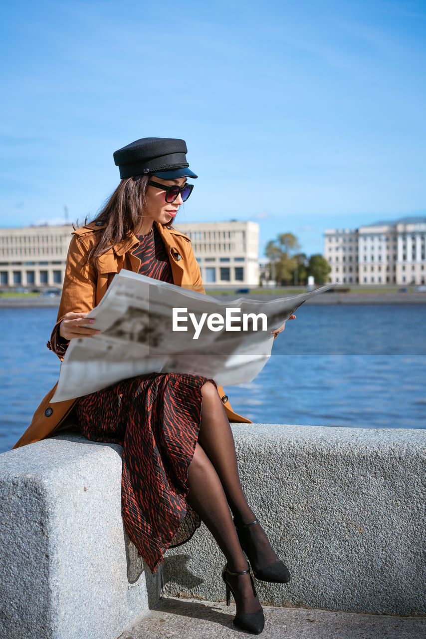 Stylish young woman reading fresh newspaper on embankment of river on sunny day