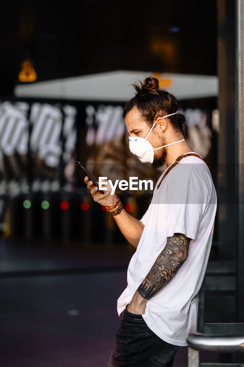 Side view of man wearing mask using mobile phone while standing outdoors