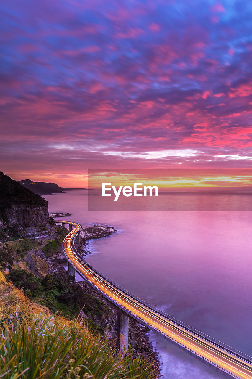Light trails on road by sea against sky at sunset