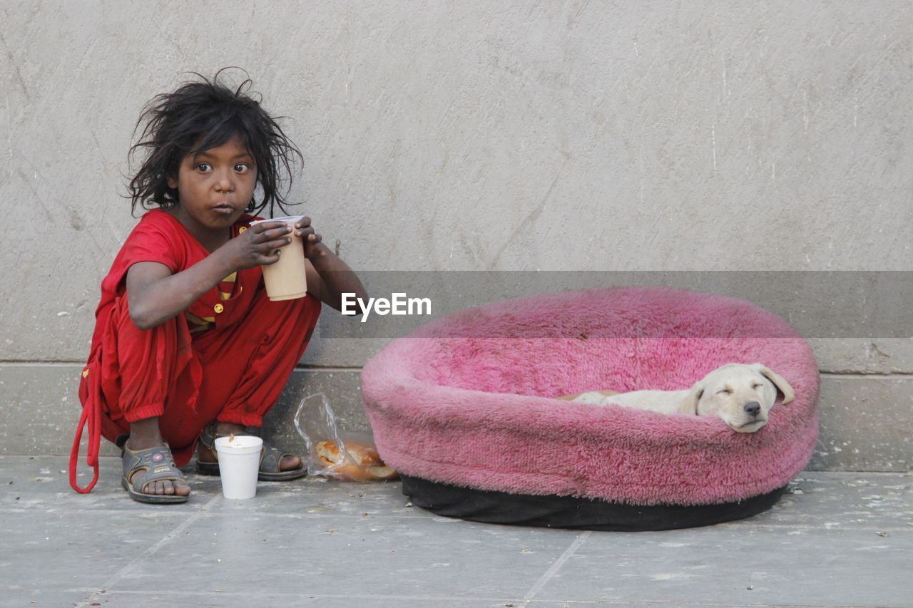 Girl holding drink looking away while sitting with puppy on sidewalk