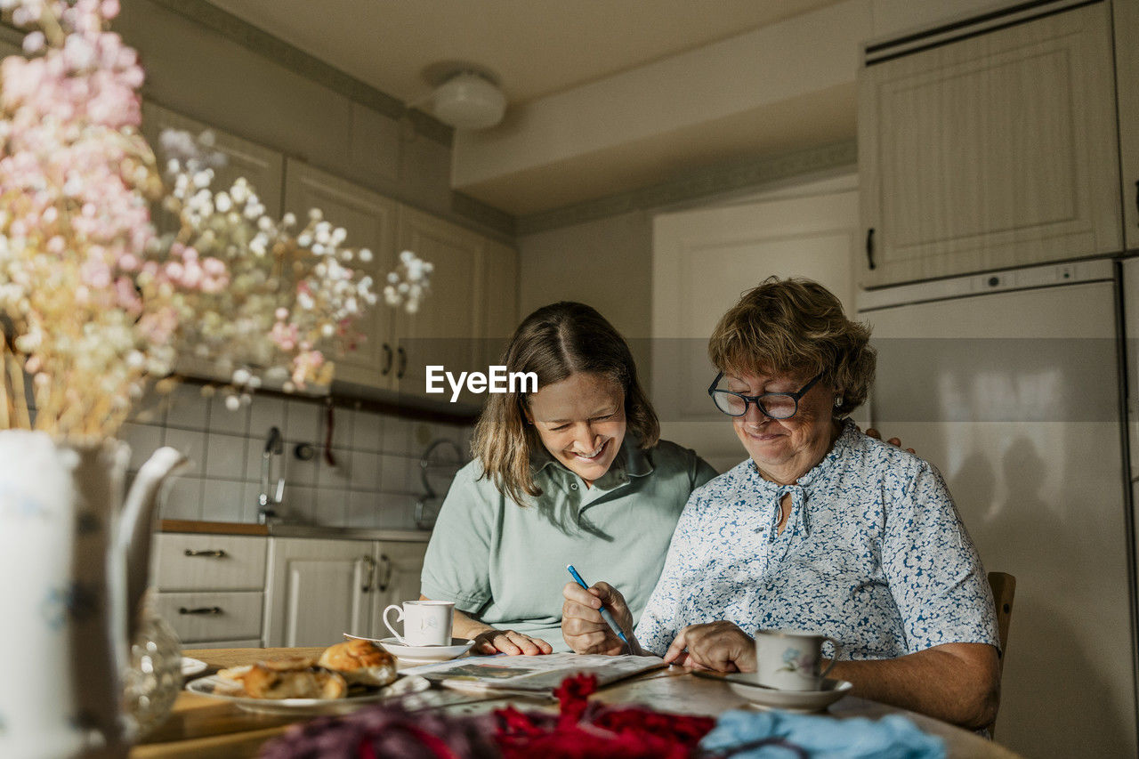 Happy senior woman and female nurse solving crossword puzzle at home
