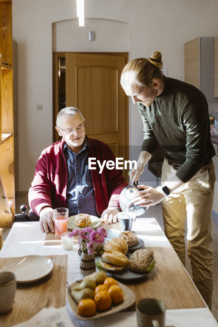 Young man pouring coffee for retired senior father with disability during breakfast at home