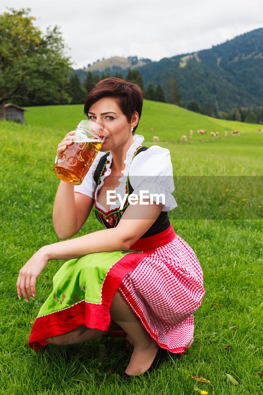 Portrait of mid adult woman drinking beer while crouching on grassy field