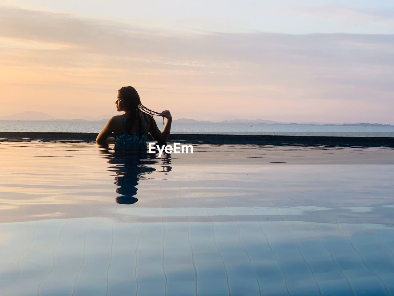 Rear view of woman in infinity pool against sky during sunset