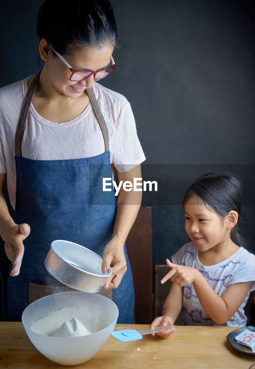 Daughter looking at mother straining flour in kitchen