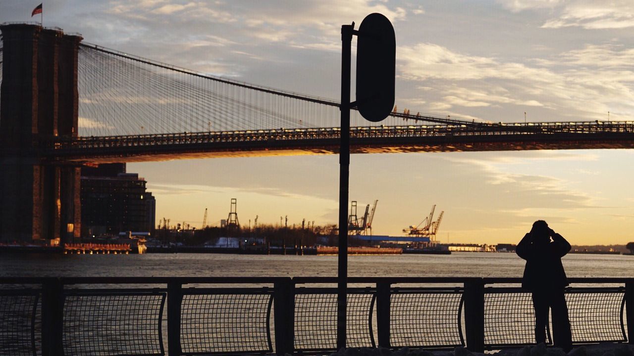 Silhouette person standing on pier looking at brooklyn bridge over east river against sky