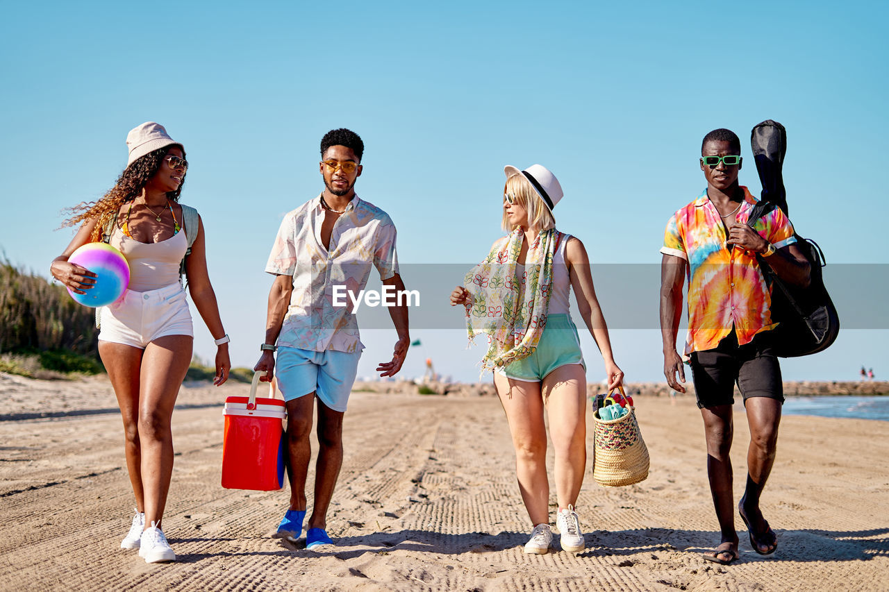 Full body multiracial men and women in sunglasses and hat strolling on sand against cloudless blue sky while spending time on beach on summer day