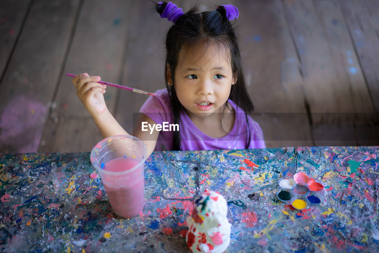 Cute girl holding paintbrush while sitting at messy table