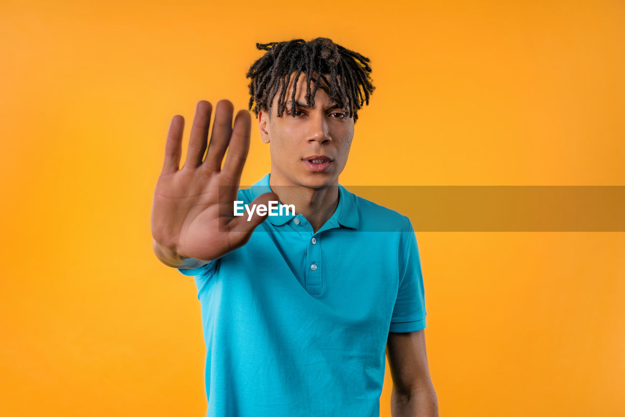 one person, colored background, studio shot, portrait, adult, orange color, waist up, yellow, men, indoors, young adult, hand, emotion, yellow background, front view, copy space, clothing, looking at camera, standing, orange background, person, gesturing, casual clothing, sign language, blue, serious, facial expression, hairstyle